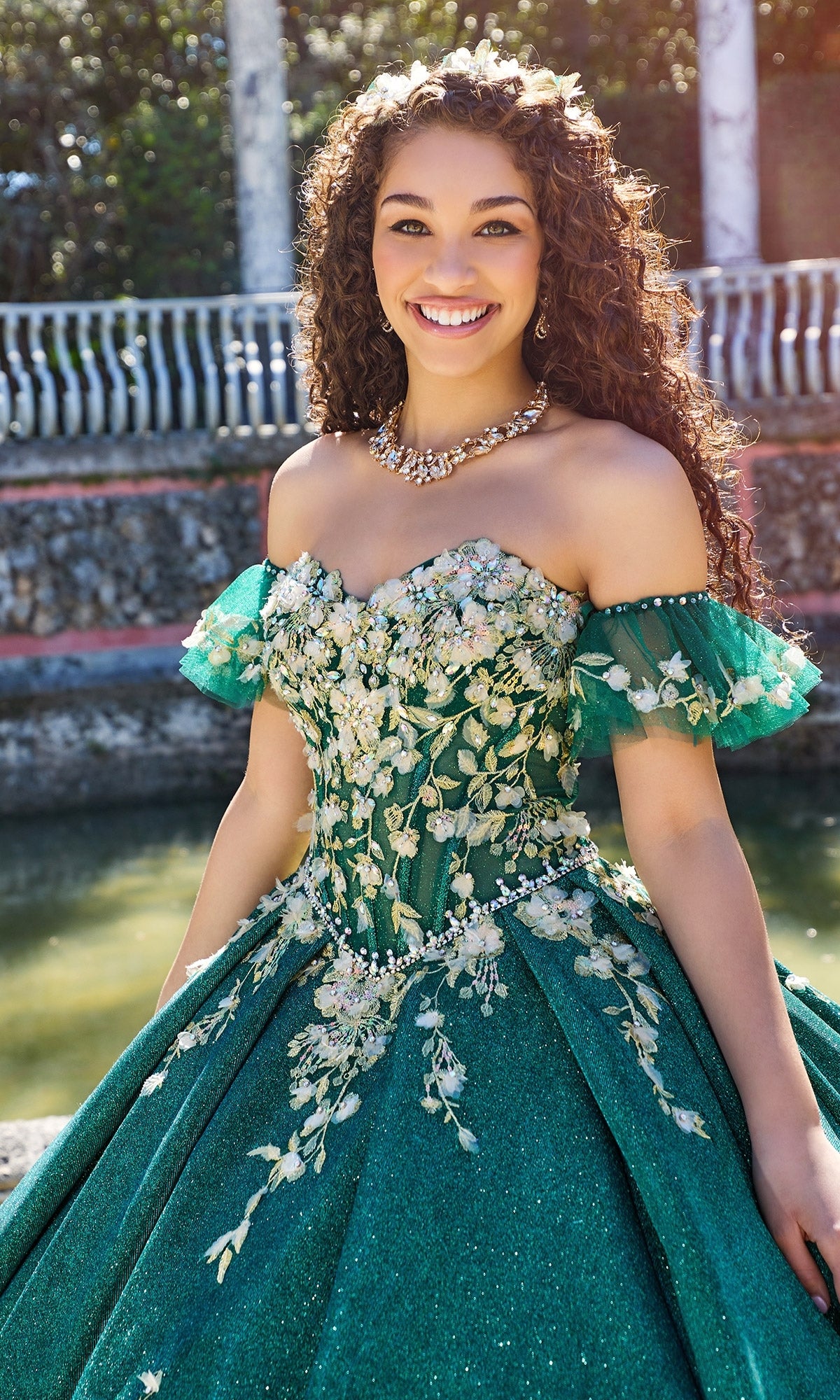 A young woman stands outdoors, smiling at the camera in the ornate Quinceanera Dress PR30134 by Princesa, featuring a green ball gown with floral embellishments and a corset bodice.